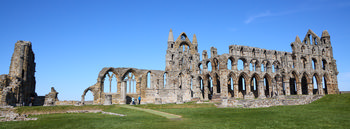 This architectural landscape photograph shows Whitby Abbey side on, capturing the imposing and intricate ruins of this historic church structure in Whitby, Yorkshire. The image was taken in the early afternoon during spring, as indicated by the bright sunlight and clear blue sky. Whitby Abbey stands prominently on the grassy hill, its gothic arches and remaining towers illustrating its former grandeur and historic significance as a religious landmark. The lush green grounds and the vivid sky enhance the details of the stonework, highlighting the Abbey’s location in Yorkshire and showcasing its role as a key heritage site in Whitby.